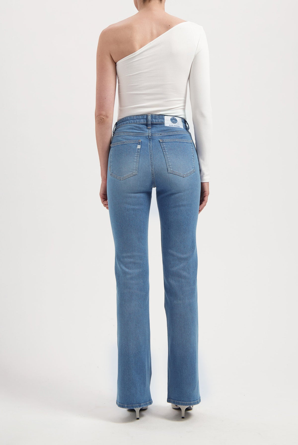 Back view of light blue, high-waisted MUD Jeans in organic cotton, worn with a white, one-shoulder long-sleeve top.