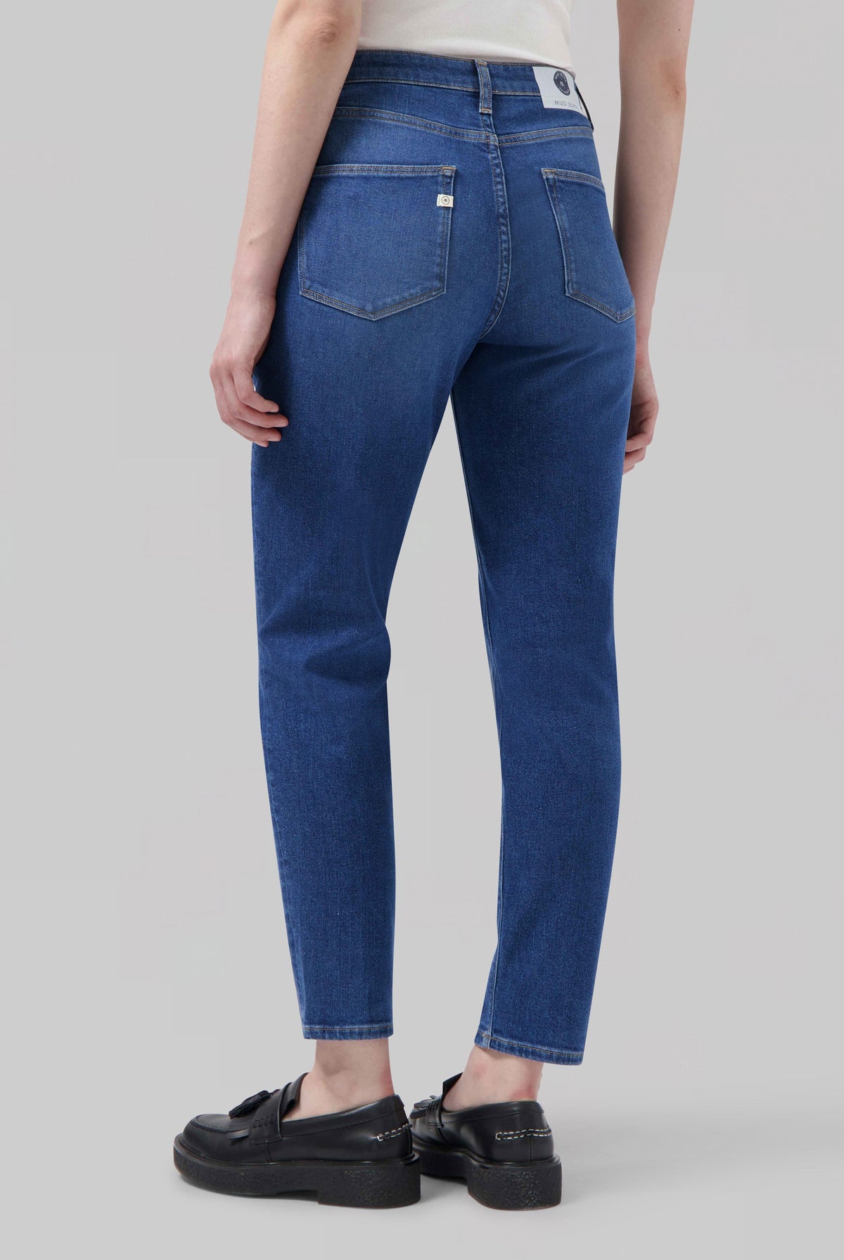 Back view of eco-friendly MUD Jeans, slim-fit, medium blue denim, shown with black loafers and a white sleeveless top