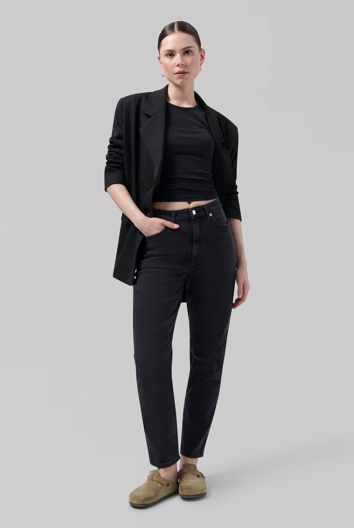 Woman wearing a black blazer over a fitted black top, paired with dark high-waisted jeans and casual tan slip-on shoes.