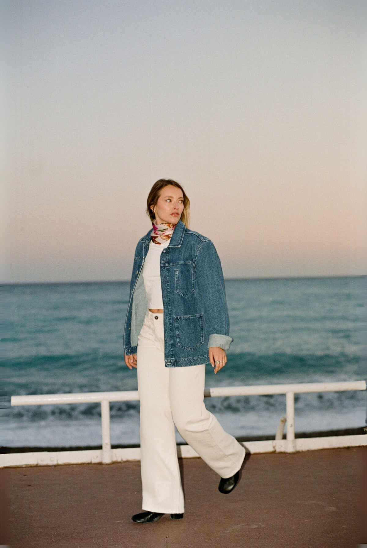 Woman in a denim jacket and white wide-leg pants standing by the ocean at sunset, wearing a colorful scarf.