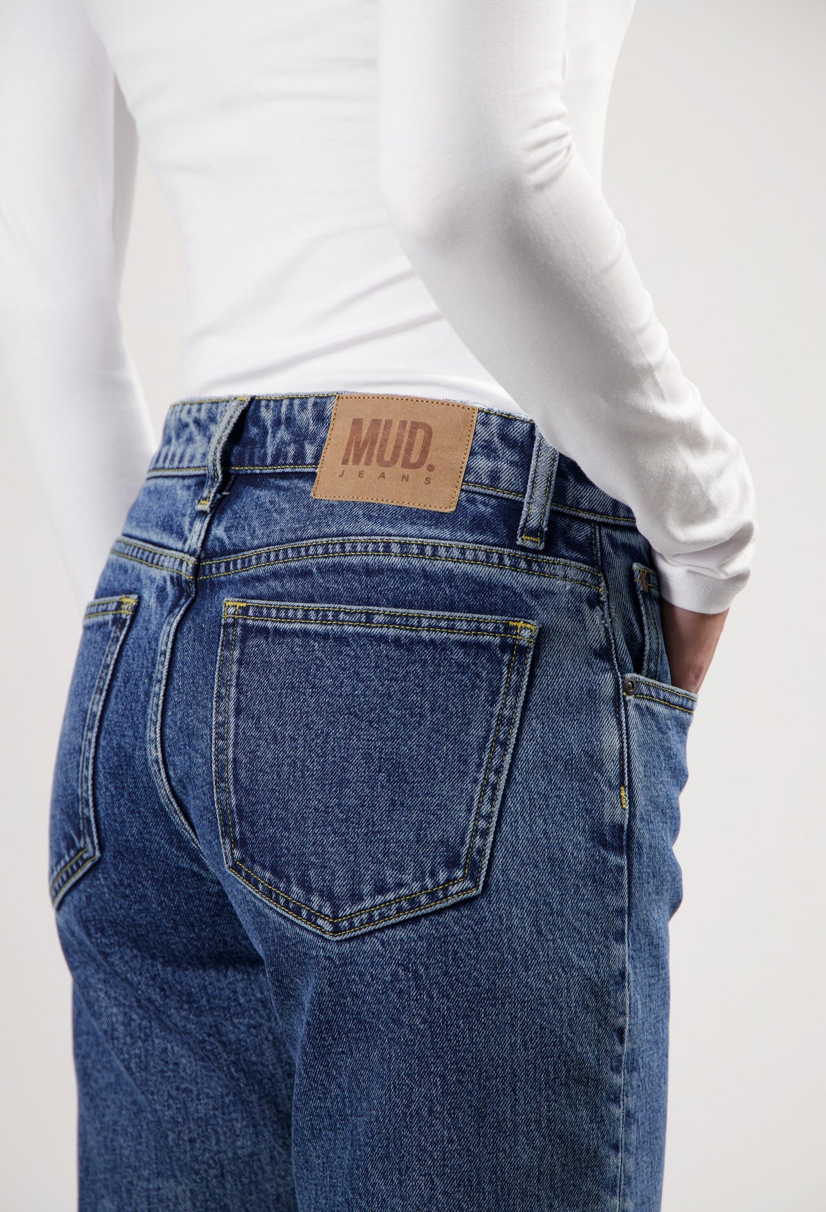 Close-up of blue MUD Jeans with a brown logo patch, worn with a fitted white long-sleeve top, showing back pocket details.