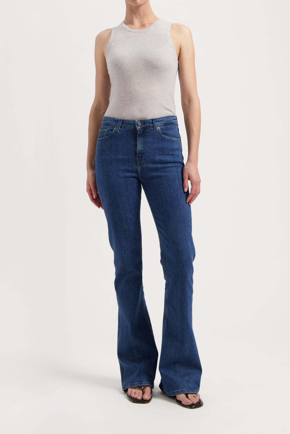 Woman wearing a light gray tank top paired with blue bootcut jeans and flip-flops, showcasing casual fashion.