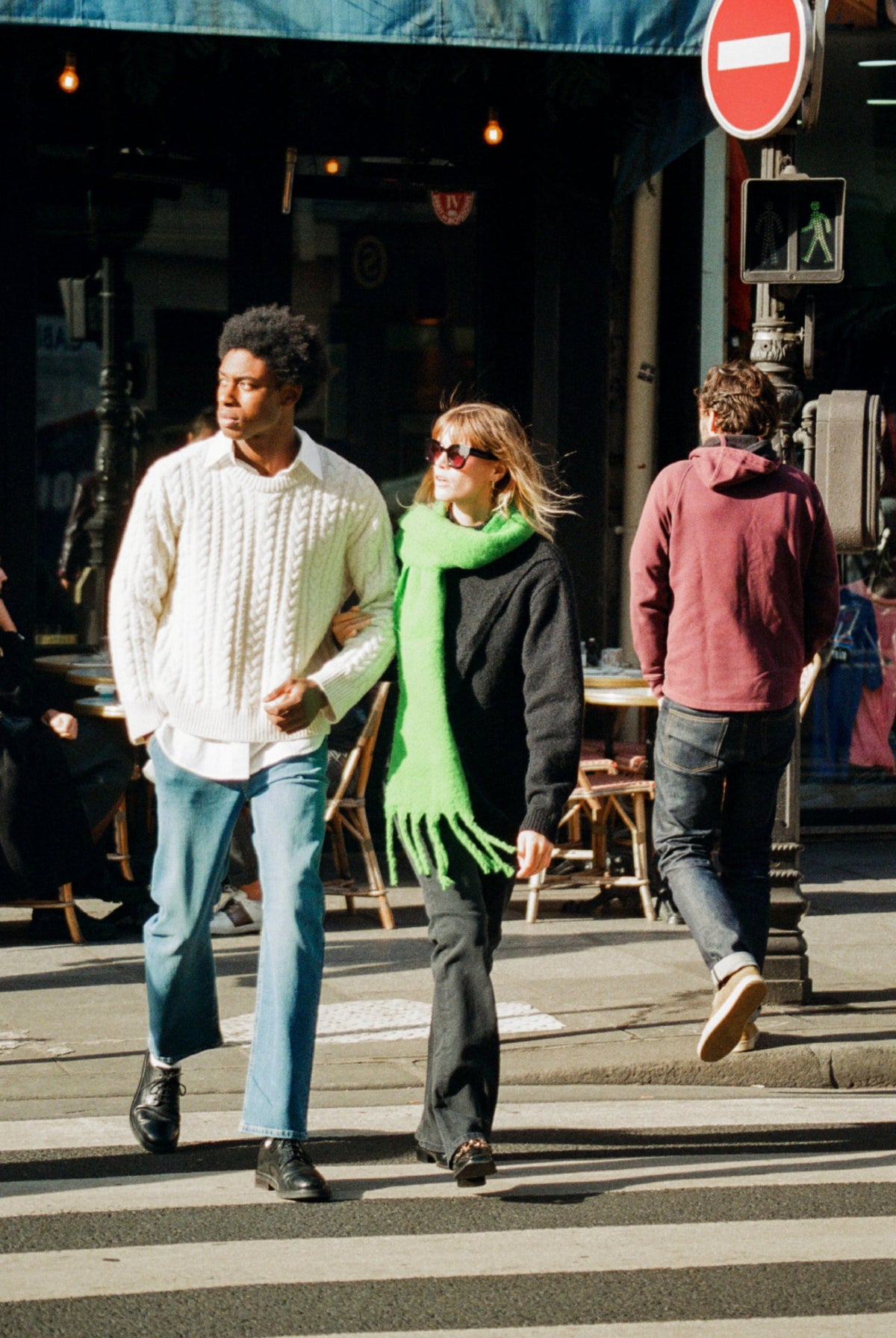 Couple crossing street wearing stylish winter outfits, including a white cable knit sweater and a bright green scarf.