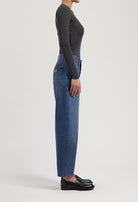 Side view of a person wearing high-waisted blue jeans and a fitted long-sleeve gray top paired with black leather loafers.