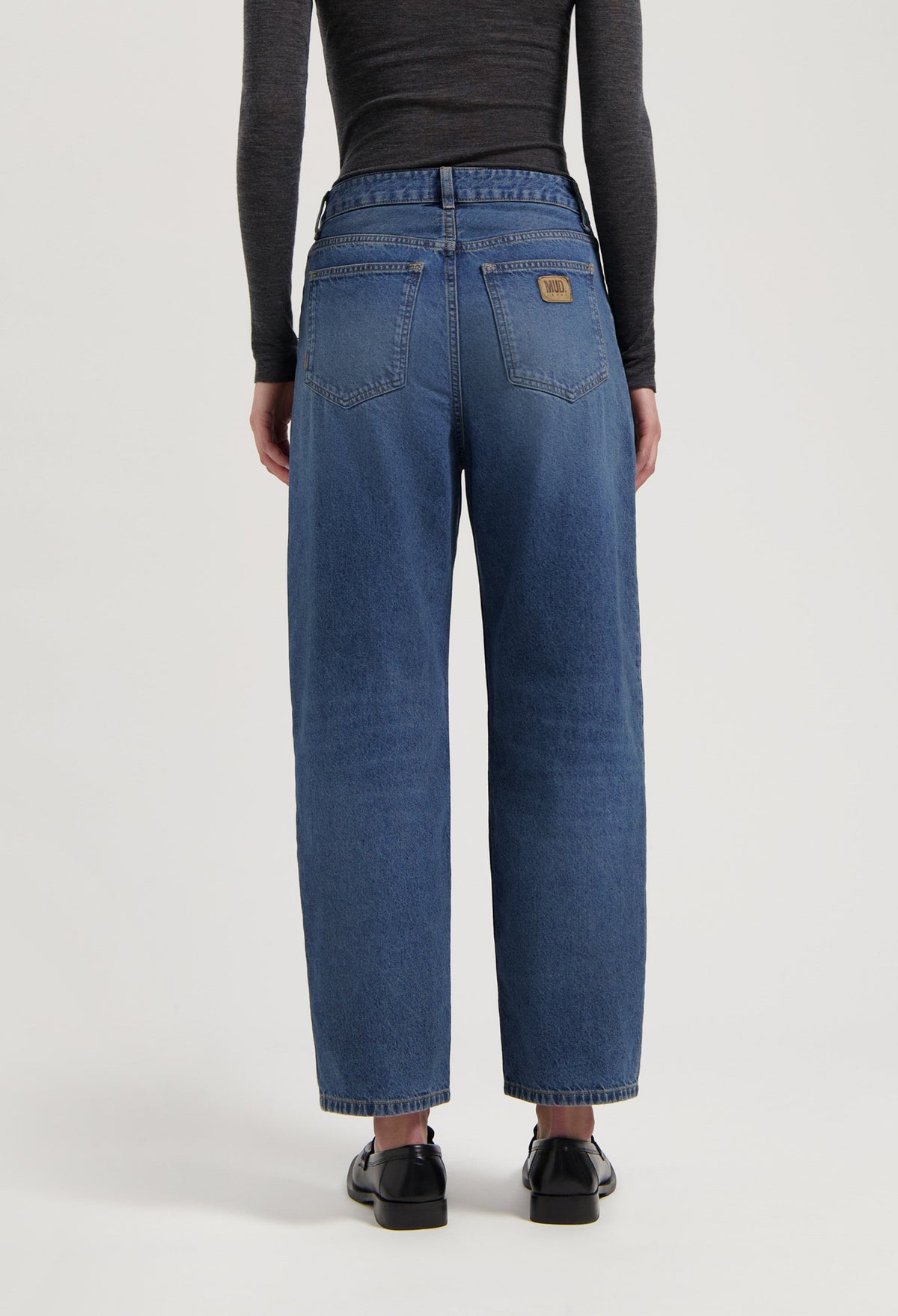 Loose-fit, mid-blue jeans by MUD Jeans made from organic cotton, styled with a dark long-sleeve top and black shoes.